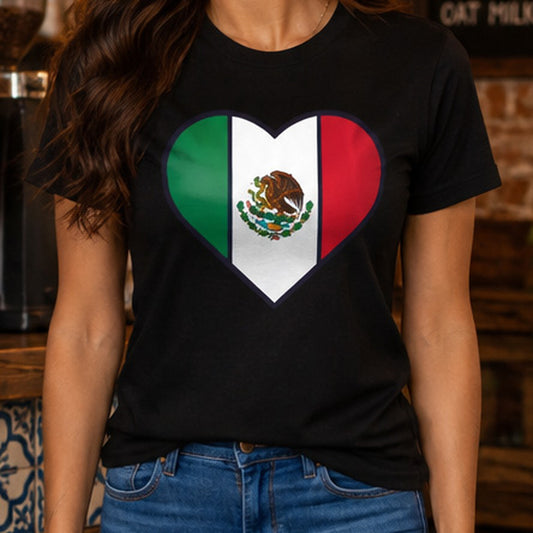 A woman wearing a black short-sleeve T-shirt with a heart-shaped Mexican flag graphic on the front, paired with blue jeans in a rustic indoor setting.